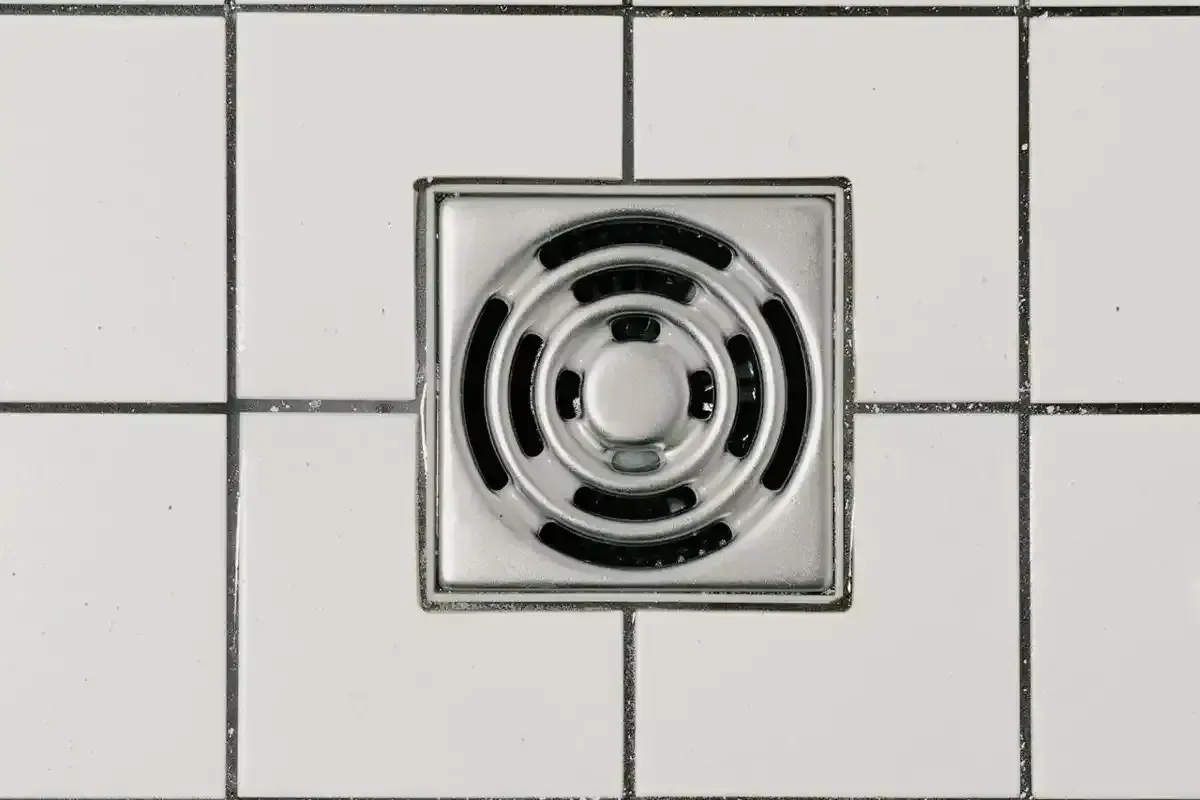 A clean, square metal shower drain with a circular grate, set in white tiled flooring with dark grout lines.
