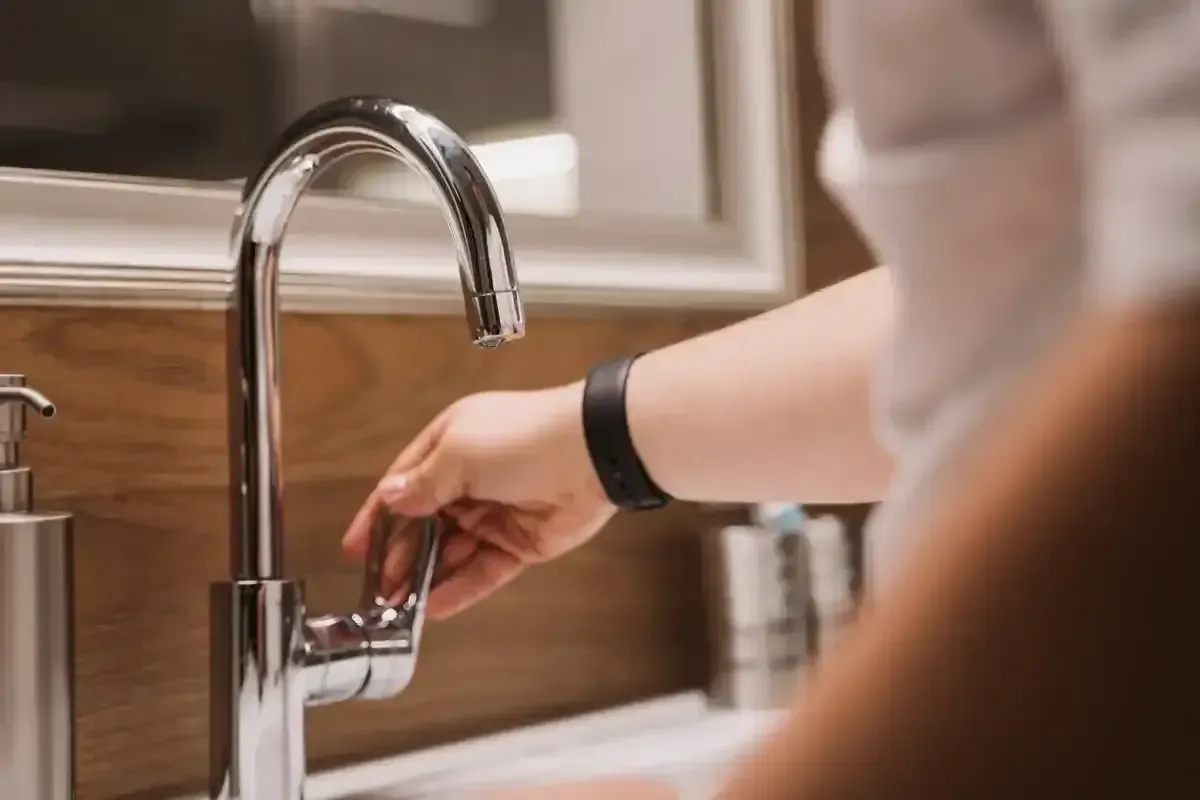 A person's hand reaching to turn on a modern chrome faucet over a white sink, with a soap dispenser nearby and a mirror reflecting blurred objects in the background.
