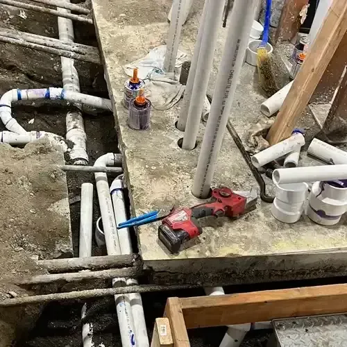 An exposed commercial plumbing installation with multiple PVC pipes, a concrete trench, and a power tool, indicating active construction or repair.