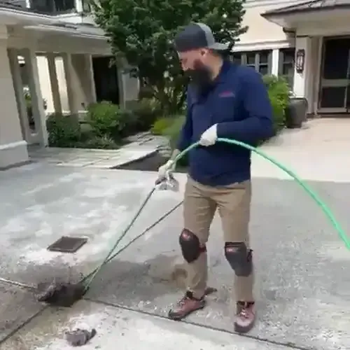 A plumber with a beard, wearing knee pads and a blue uniform, operating a hydro-jetting hose to clear a drain on a paved driveway in front of a house.