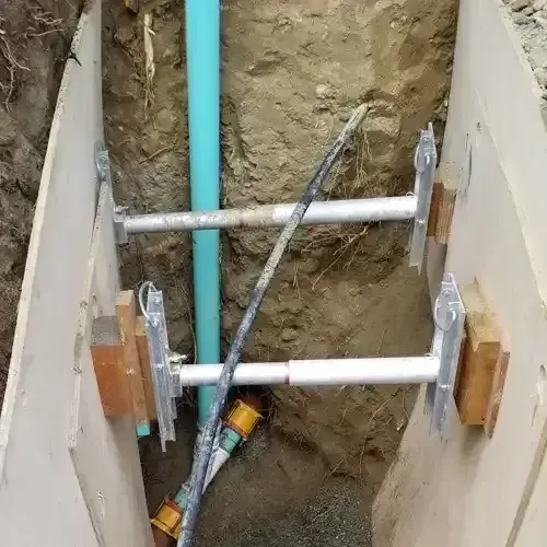 A trench with shoring equipment bracing the walls, revealing a light blue sewer pipe and other utility lines underground, indicating a repair in progress.