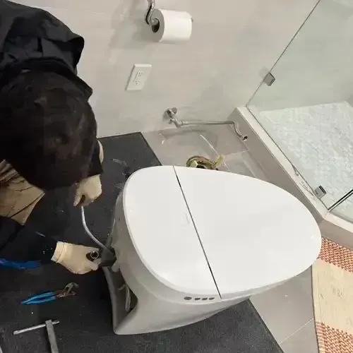 A plumber with work gloves is installing a modern, white, bidet-style toilet in a bathroom with tiled walls and a glass shower.