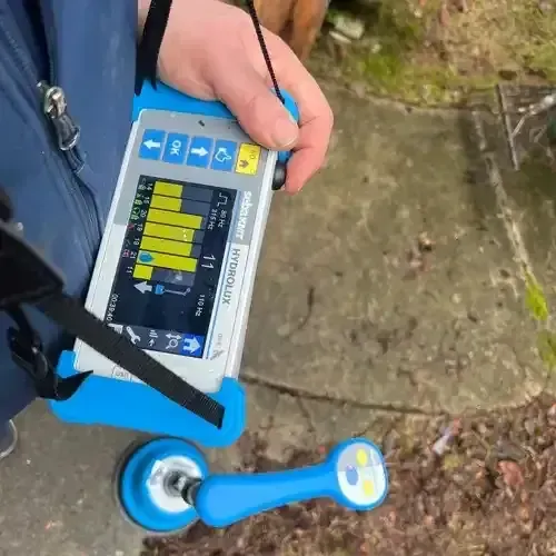 A person holding a blue and white SebaKMT Hydrolux water leak detection device with a ground microphone on a paved outdoor surface.